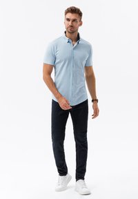 Camisa de botones de manga corta azul claro con cuello amplio, combinada con jeans oscuros y zapatillas blancas. Textura de tela suave y diseño ajustado.