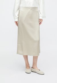 Μίντι φούστα από ελαφρύ μπεζ σατέν με ευθεία γραμμή, διαθέτοντας απαλή υφή και μίνιμαλ σχεδιασμό. Φορεμένη με μαλακά, ουδέτερα φλατ παπούτσια.