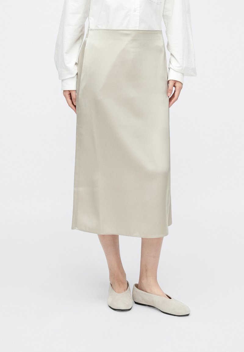 Μίντι φούστα από ελαφρύ μπεζ σατέν με ευθεία γραμμή, διαθέτοντας απαλή υφή και μίνιμαλ σχεδιασμό. Φορεμένη με μαλακά, ουδέτερα φλατ παπούτσια.