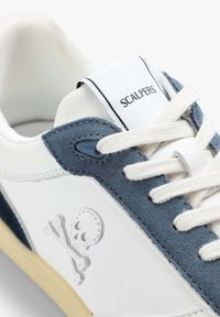 Weiße Sneaker mit blauen Wildlederakzenten, featuring ein Totenkopf- und Kreuzbones-Emblem, Stoffsenkel und einer gepolsterten Zunge, die mit "SCALPERS" beschriftet ist.