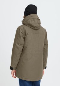 Olivenfarget parka med hette, justerbare mansjetter og rett snitt. Laget av teksturert stoff med en glatt overflate.