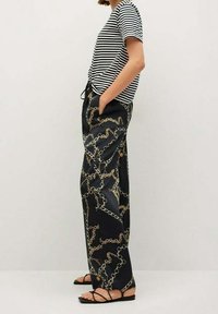 Persoon in zwarte wijde broek met gouden kettingprint, zwart-wit gestreept T-shirt met korte mouwen en zwarte platte sandalen met bandjes.