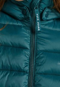 Tealfärgad puffjacka med en slät, glänsande yta, med en mittdragkedja och logotypetikett. Vadderad design med tryckknappar på kragen.