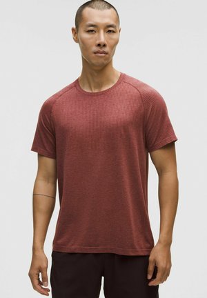 Piros textúrájú, rövid ujjú póló kerek nyakkivágással és raglan ujjakkal, sötét bermudával viselve, semleges háttér előtt.