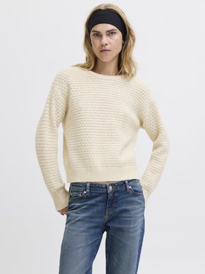 Krämfärgad stickad tröja med ett strukturerat mönster och ribbad fåll, i kombination med mörkblå denimjeans. Modellen bär ett svart hårband.