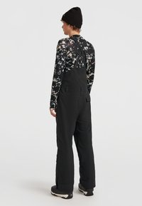 Zwarte overall met verstelbare bandjes, voorzien van een zijzak en een relaxte pasvorm. Het model draagt een gestreept shirt met lange mouwen en een muts.