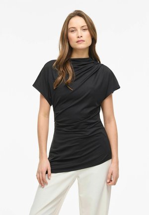 Donna con capelli lunghi e castani indossa una maglietta nera aderente a maniche corte e pantaloni bianchi larghi, in piedi davanti a uno sfondo bianco semplice.