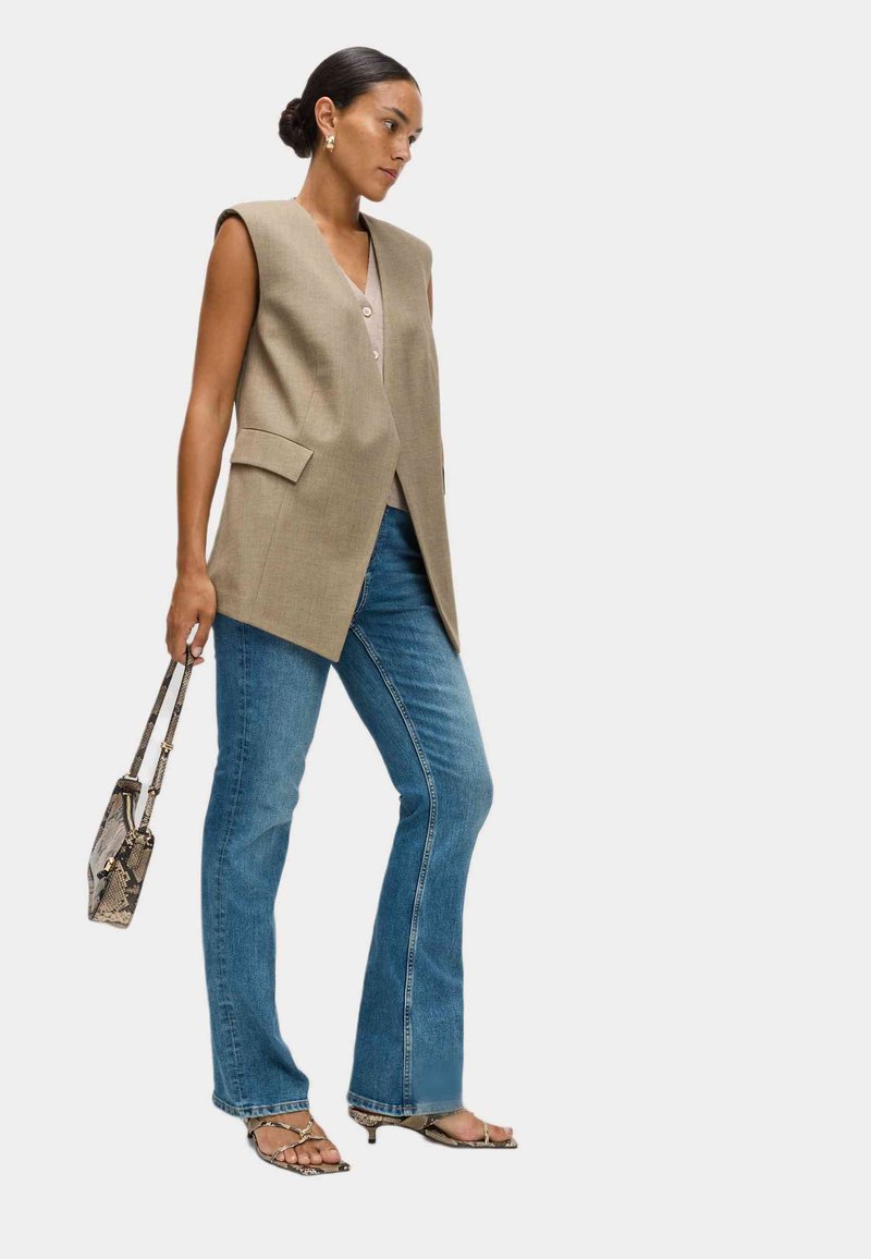 Blazer beige senza maniche con due tasche anteriori, abbinato a jeans a zampa d'elefante blu. Il modello tiene in mano una piccola borsa in stampa pitone e indossa sandali.
