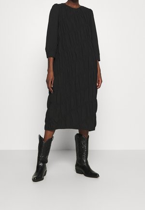 Vestido negro con diseño texturizado y plisado, de mangas tres cuartos, combinado con botas vaqueras negras con costuras decorativas.