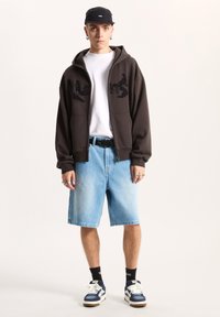 Sudadera marrón con cremallera y diseño negro bordado, camiseta blanca, pantalones cortos de mezclilla azul claro, cinturón negro y zapatillas azules. Gorra negra y anillos.