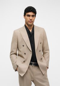 Homme aux cheveux noirs bouclés portant un costume croisé beige et une chemise noire, les mains dans les poches, debout devant un fond neutre.