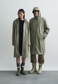 To personer har på seg olivengrønne regnfrakker, knelange gummistøvler og lysebrune bukser. Frakkene har hetter og snøresnor i livet.