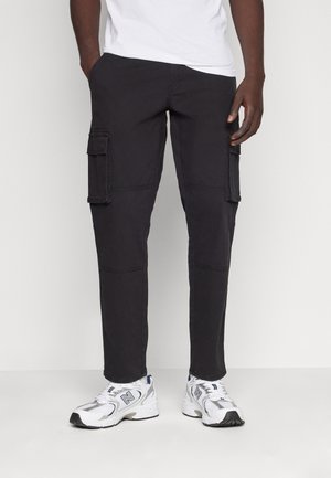 Man wearing black cargo pants with flap pockets and white athletic sneakers standing against a plain light background.