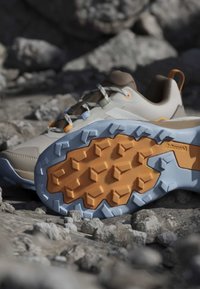 Chaussure de randonnée avec une tige beige et une semelle robuste bleu-orange reposant sur un terrain rocheux, mettant en avant le motif d'adhérence et les détails du lacet.