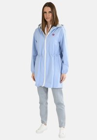 Veste imperméable bleu clair avec une fermeture éclair, une taille élastique, des poches et une capuche. Associée à un jean denim clair et des baskets blanches.