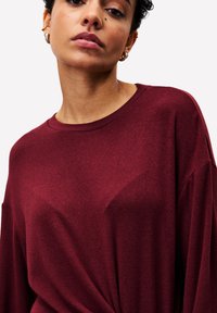 Femme portant des boucles d'oreilles en or et un haut à manches longues maroon avec un col rond devant un fond uni.