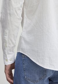 Camisa de lino blanca de mangas largas y puños con botones, combinada con jeans de denim azul. La camisa tiene un dobladillo curvado y una textura de tela visible.