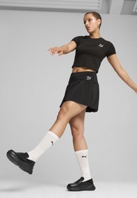 Černé zkrácené tričko s logem Puma, černá skladaná sukně, bílé žebrované ponožky s logem Puma a černé chunky boty.