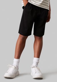 Pantaloni corti neri con tasche laterali, realizzati in materiale morbido e con una vestibilità comoda, abbinati a sneakers bianche e calze bianche a coste.