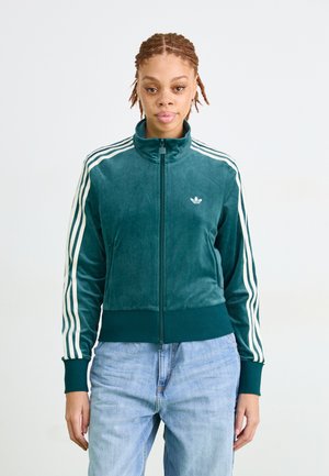 Person with braided hair wearing a teal zip-up Adidas track jacket with white stripes and light blue jeans against a white background.