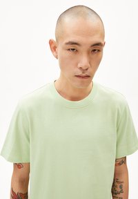 Hellgrünes Baumwoll-T-Shirt, Rundhalsausschnitt, kurze Ärmel, glatte Textur, mit sichtbaren Tattoos auf dem Arm der Person.