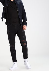 Person som bär svarta trasiga jeans, svart skjorta, mörkblå jacka, vita strumpor och vita sneakers som står mot en enfärgad bakgrund.