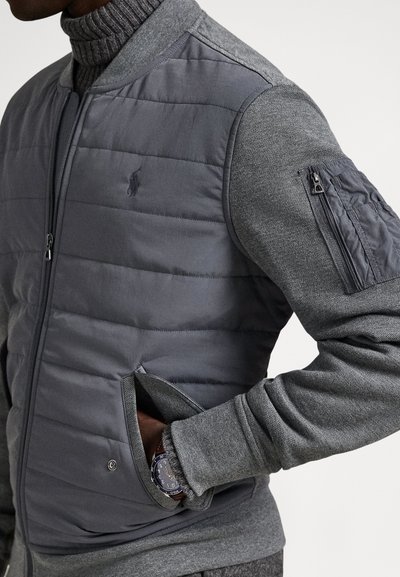 Mężczyzna w szarej pikowanej kurtce z dzianinowymi rękawami i zamykaną kieszenią na górnym ramieniu, ręka w bocznej kieszeni, pokazująca zegarek na nadgarstku.