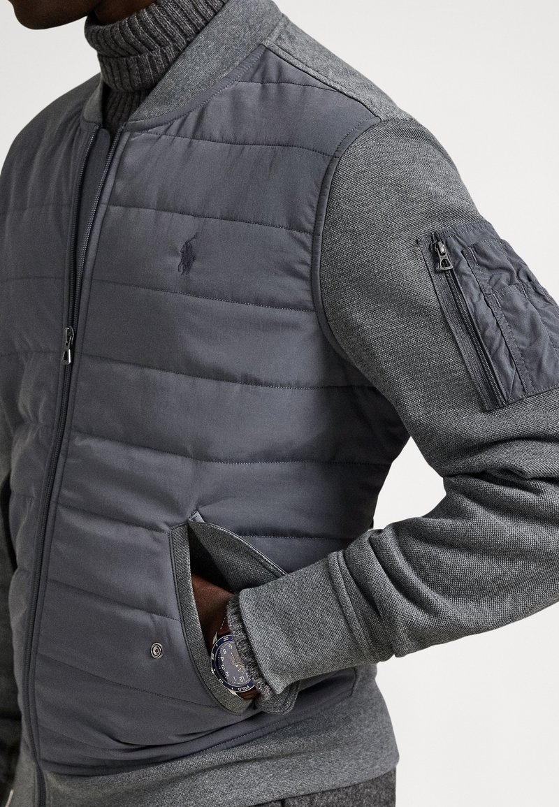 Homme portant une veste matelassée grise avec des manches en tricot et une poche zippée sur le haut du bras, main dans la poche latérale montrant une montre-bracelet.