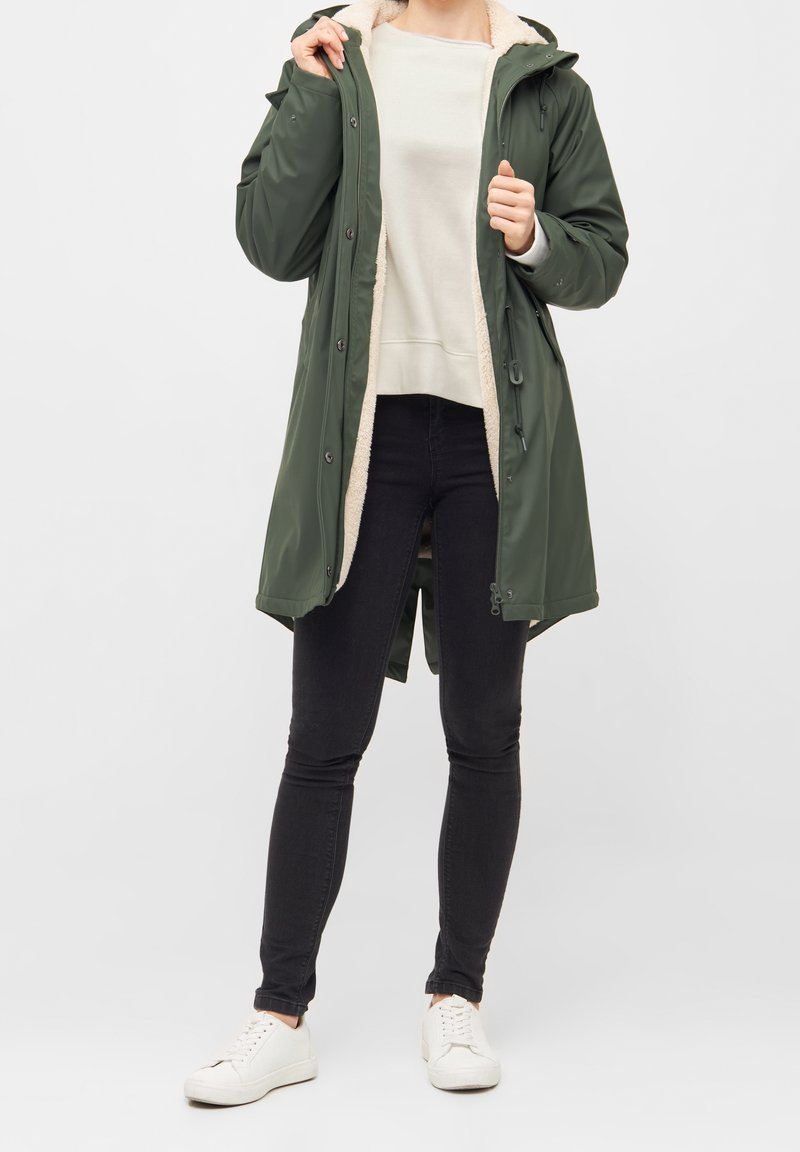 Parka verde oliva con una morbida fodera beige, cappuccio, chiusura a bottoni e cerniera, vita regolabile con coulisse e tasche laterali. Indossati jeans e sneakers.