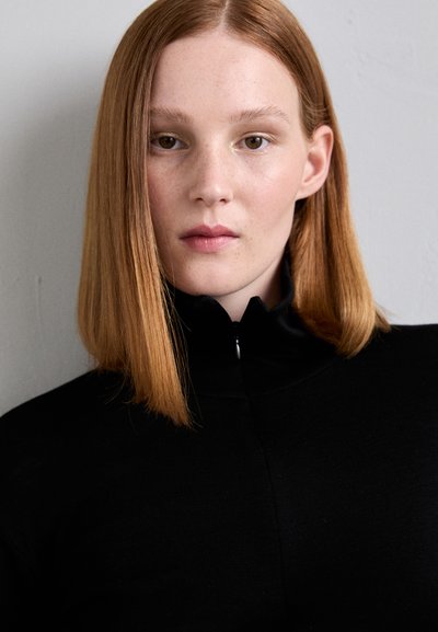 Musta vetoketjullinen toppi, jossa on korkea kaulus, tiukka muotoilu ja pehmeä pinta. Mallilla on suorat, hartioille ulottuvat punaiset hiukset.