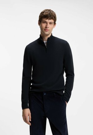 Junger Mann, der einen schwarzen Pullover mit Reißverschluss und dunkle Hose trägt, steht mit einer Hand in der Tasche vor einem schlichten weißen Hintergrund.