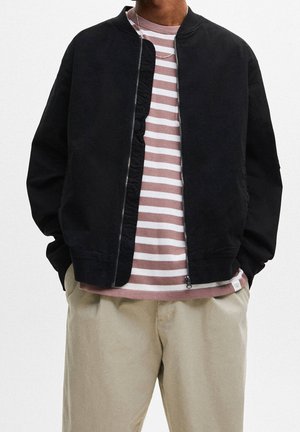 Person med hænder i lommerne iført sort jakke med lynlås over en pink og hvidstribet skjorte og beige bukser mod en ensfarvet baggrund.