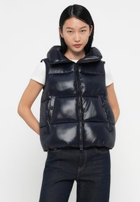 Marine puffer vest met hoge kraag, glanzende afwerking, ritssluiting aan de voorkant en twee knoopzakken, gedragen over een witte blouse.