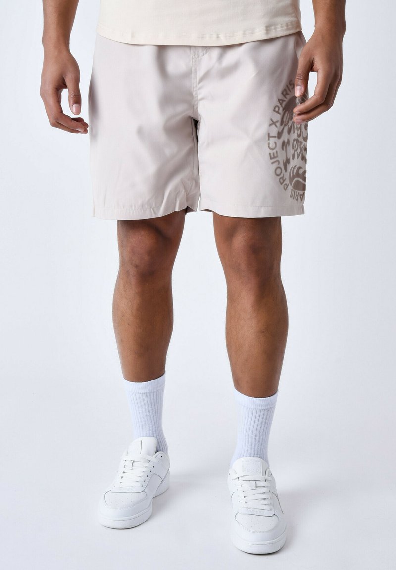Bañador color beige con una textura suave, que presenta un gráfico circular en marrón en la pierna izquierda. Se lleva con calcetines deportivos blancos y zapatillas.