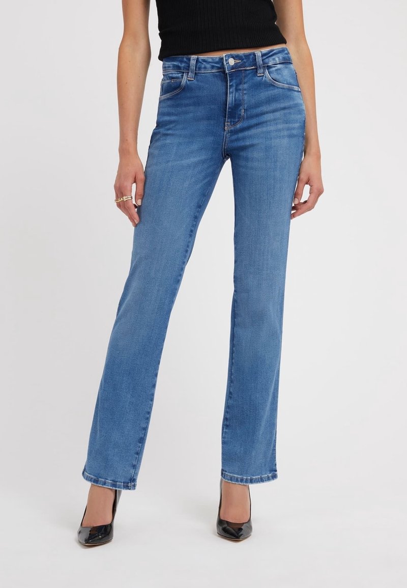 Femme portant un jean droit bleu, un haut noir sans manches, des talons aiguilles noirs pointus et des bagues en or à la main gauche.