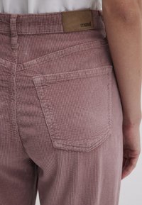Rosa Cordhosen weisen eine hohe Taille, vertikale Rippungen und ein Leder-Logopatch auf. Eine Rückentasche ist sichtbar und zeigt den strukturierten Stoff.
