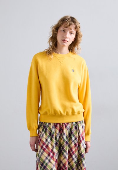 Sudadera de algodón amarilla con puños y dobladillo acanalados, que presenta un pequeño logo azul. Combinada con una falda de cuadros multicolor.