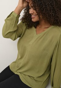 Blusa verde oliva con scollatura a V e tessuto a trama, dotata di maniche lunghe e voluminose e una vestibilità rilassata, abbinata a pantaloni neri.