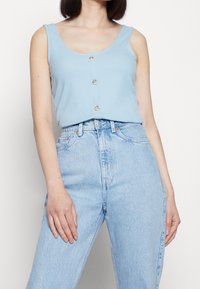 Light blue ribbed tank top featuring a scoop neckline and three buttons down the front, paired with high-waisted light denim jeans.