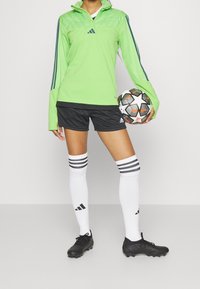 Zielona sportowa bluzka z długim rękawem zdobiona pikowanym wzorem, czarne szorty, białe skarpetki z trzema czarnymi paskami oraz czarne korki piłkarskie, trzymająca piłkę nożną.