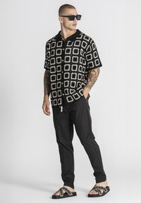 Mann mit Tätowierungen, der ein kurzärmeliges Hemd mit schwarz-weißem Schachbrettmuster, schwarze Hose, Sonnenbrille und beige Sandalen mit schwarzen Riemen trägt.