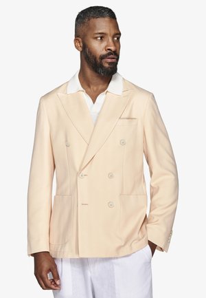 Mężczyzna z brodą, ubrany w beżową dwurzędową marynarkę i białą koszulę, patrzący w prawo, z jedną ręką w kieszeni.