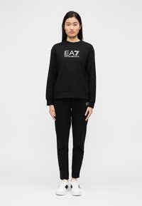 Μαύρο φούτερ με το λογότυπο EA7 και μαύρο παντελόνι που ταιριάζει. Λαιμόκοψη σε στυλ crew και μακριά μανίκια, συνδυασμένο με λευκά αθλητικά παπούτσια για αντίθεση.