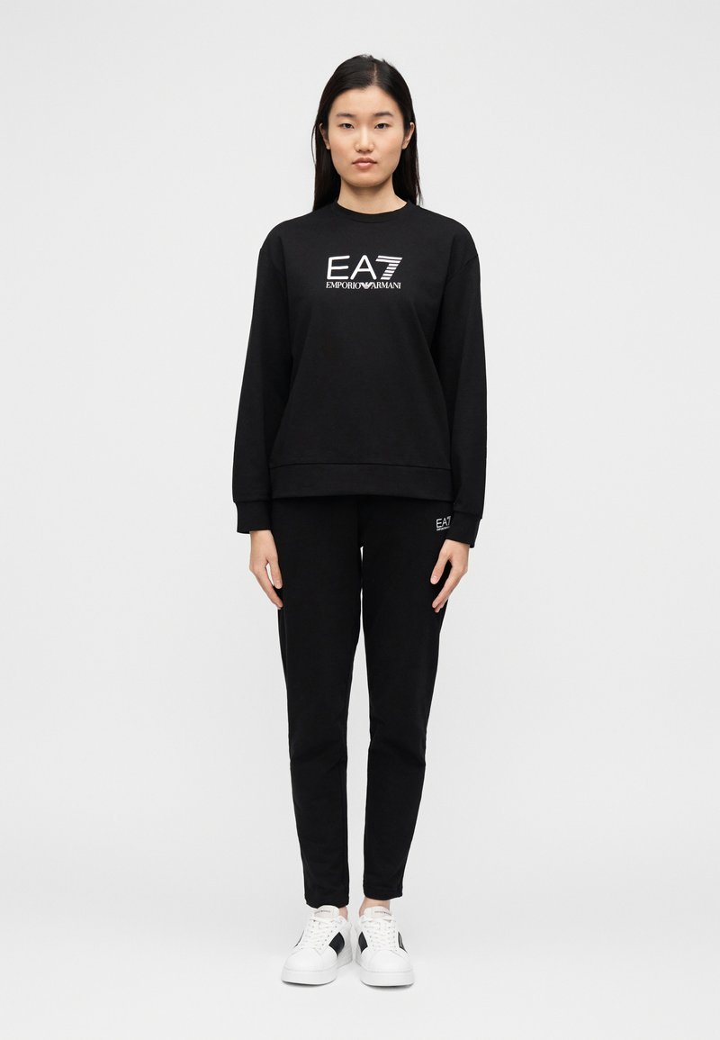 Μαύρο φούτερ με το λογότυπο EA7 και μαύρο παντελόνι που ταιριάζει. Λαιμόκοψη σε στυλ crew και μακριά μανίκια, συνδυασμένο με λευκά αθλητικά παπούτσια για αντίθεση.