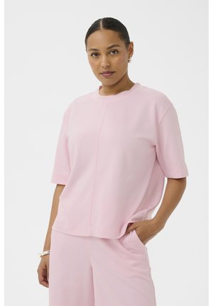 Femme aux cheveux bruns tirés en arrière, portant un haut rose clair à manches courtes et un pantalon assorti, debout avec une main dans la poche, sur fond blanc.