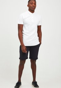 White polo shirt with short sleeves, paired with navy rolled-up shorts. Black sneakers complete the casual look. Smooth cotton texture.