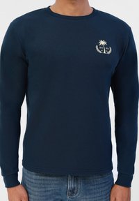 Sweatshirt bleu marine à manches longues en coton, arborant un petit logo de palmier et un texte crème, avec un col rond et des poignets côtelés.