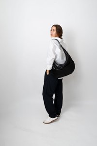 Mulher com uma camisa branca e calças risca de giz, vestindo ténis brancos e carregando uma grande mala preta de ombro, de pé contra um fundo branco liso.