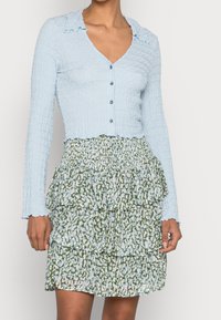 Cardigan long bleu clair, texturé, à manches longues avec un col froncé et des boutons. Associé à une jupe à volants à motifs verts avec des accents bleus.