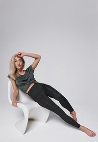 Zwarte, aansluitende leggings en een donkergroene, cropped t-shirt met een knoopdetail, gedragen terwijl zittend op een witte moderne stoel.